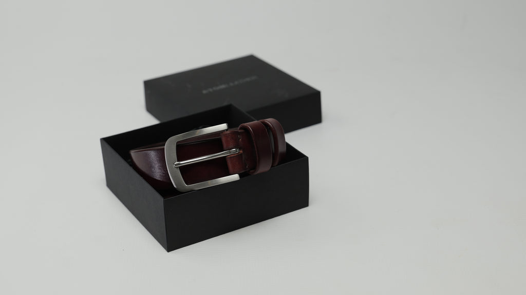 A mahogany burgundy leather belt with a stainless steel buckle, presented inside an open black gift box with its lid placed behind it on a light gray background.