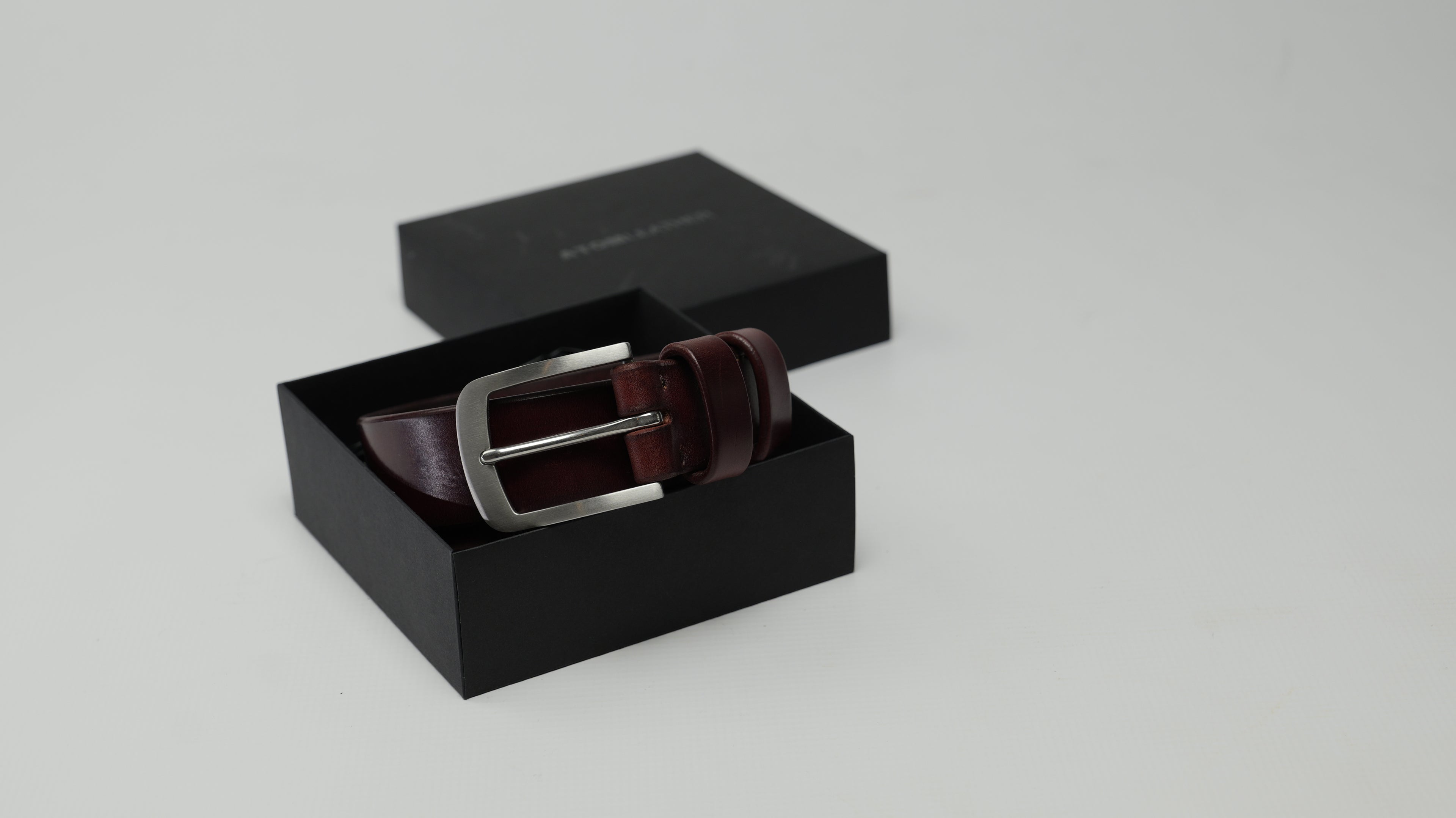 A mahogany burgundy leather belt with a stainless steel buckle, presented inside an open black gift box with its lid placed behind it on a light gray background.
