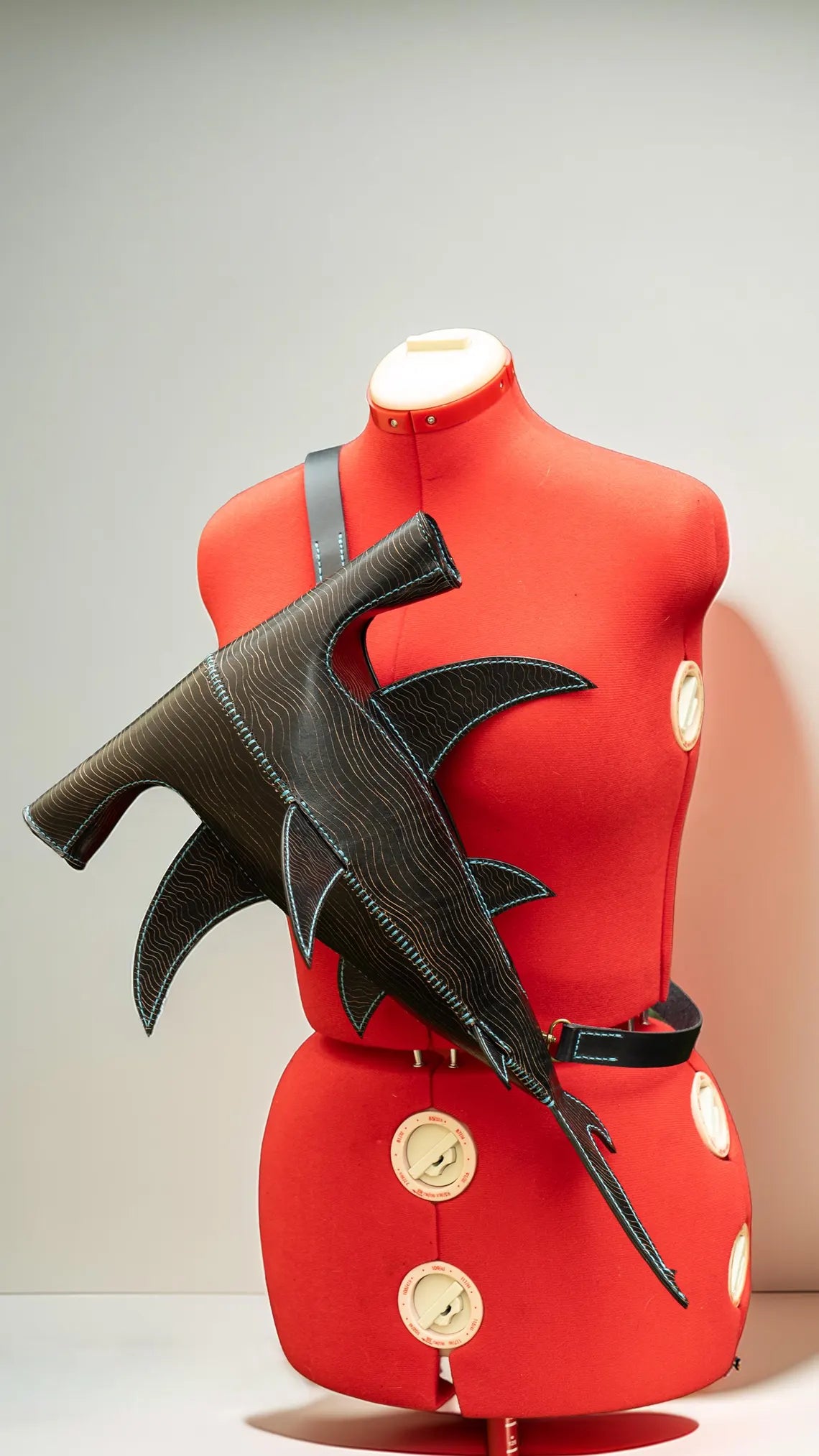 A black textured leather shoulder bag with a shark fin design, displayed on a red mannequin torso against a light gray background. Made by ATOM Leather.