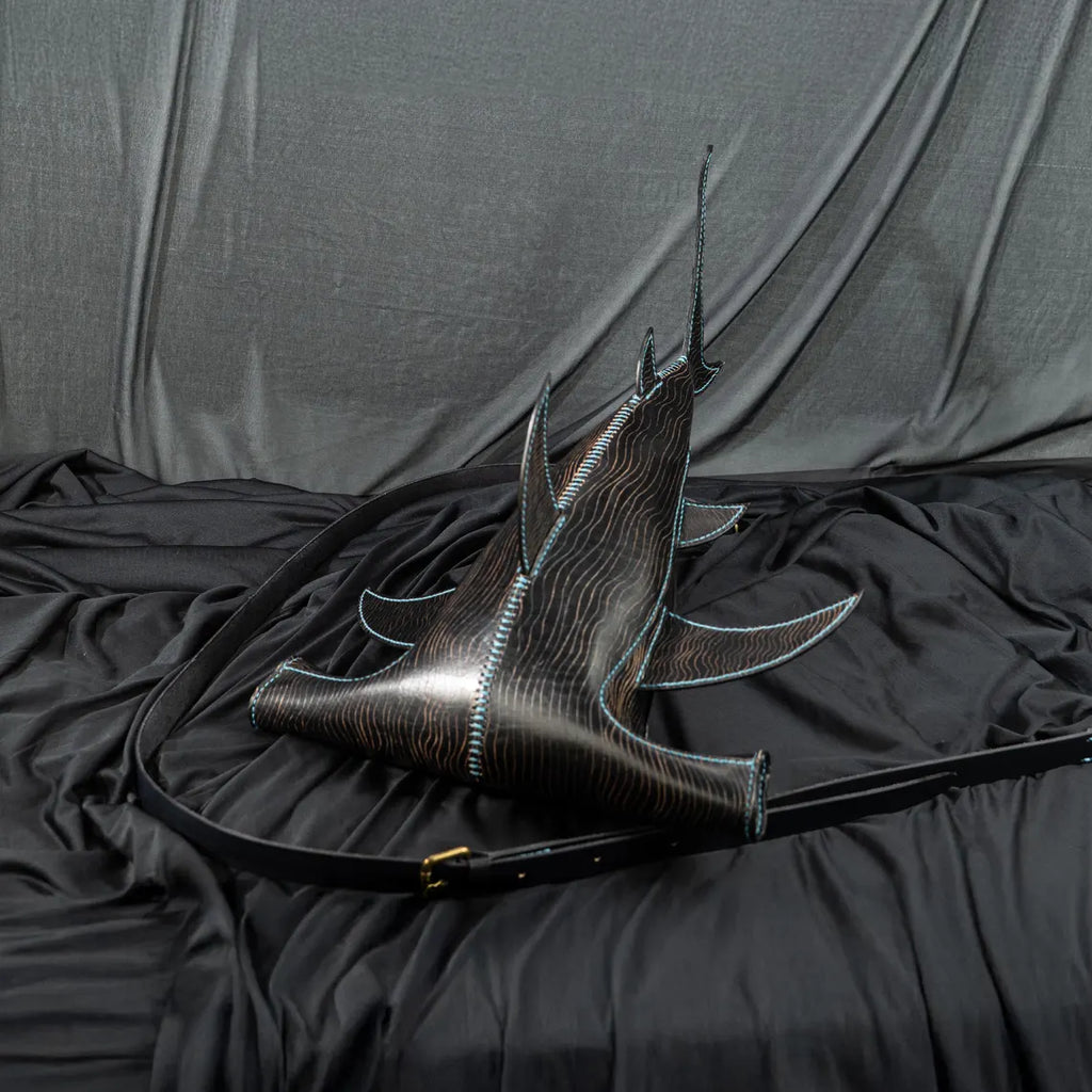 A black leather shoulder bag with a sculptural, fish-inspired silhouette, featuring a glossy dark wood-grain texture, light blue contrast stitching, and a long black strap, displayed on a black fabric backdrop. Made by ATOM Leather.