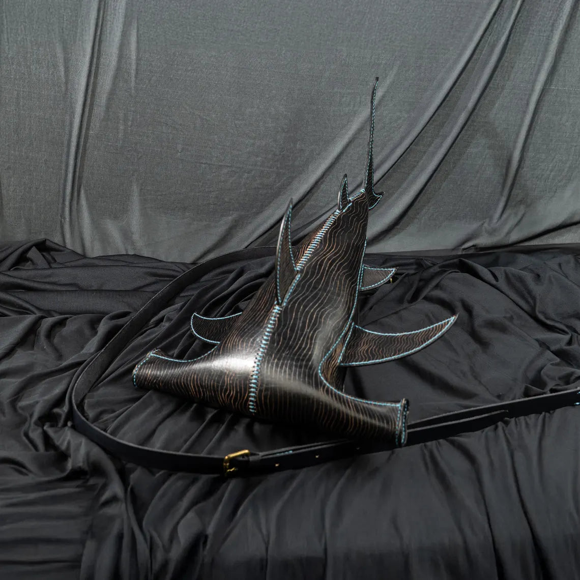 A black leather shoulder bag with a sculptural, fish-inspired silhouette, featuring a glossy dark wood-grain texture, light blue contrast stitching, and a long black strap, displayed on a black fabric backdrop. Made by ATOM Leather.