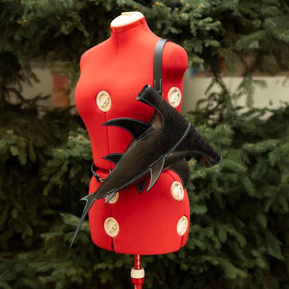 A black leather shark-shaped shoulder bag with a textured scale pattern and silver hardware, displayed on a red dress form mannequin with a blurred green pine tree background. Made by ATOM Leather.