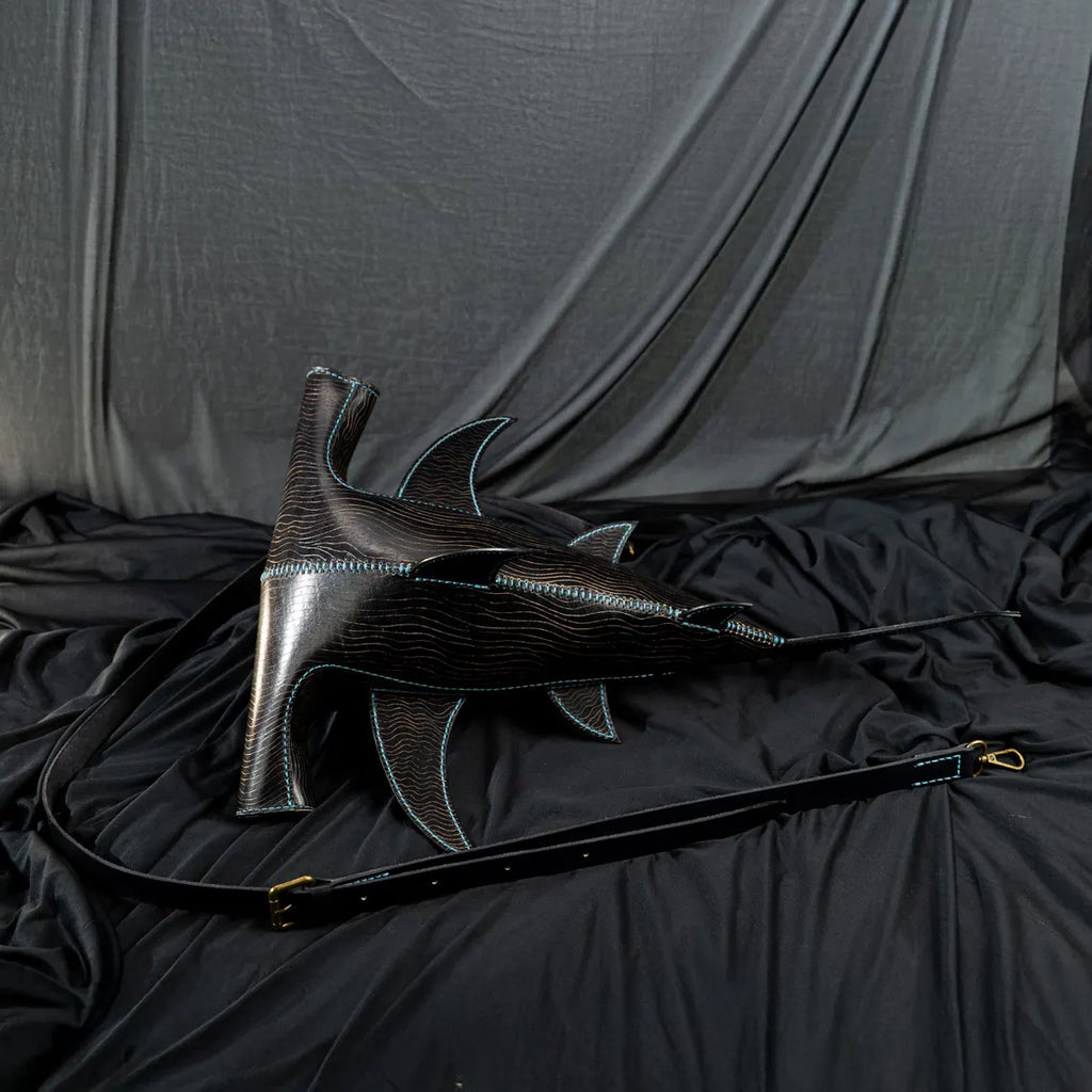 A black shark-shaped leather shoulder bag with light blue contrast stitching and a long black strap, displayed on a black fabric backdrop. Made by ATOM Leather.