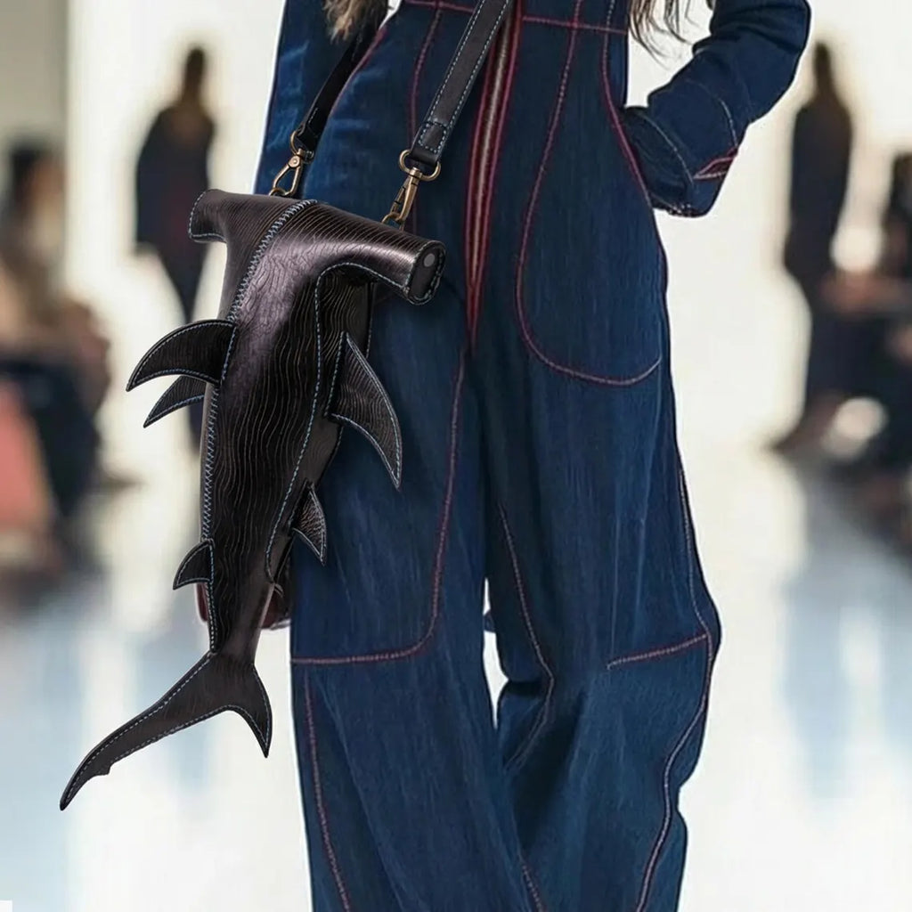 A person wearing dark blue denim overalls with red contrast stitching carries a dark brown shark-shaped leather shoulder bag in a blurred indoor. setting. Made by ATOM Leather.
