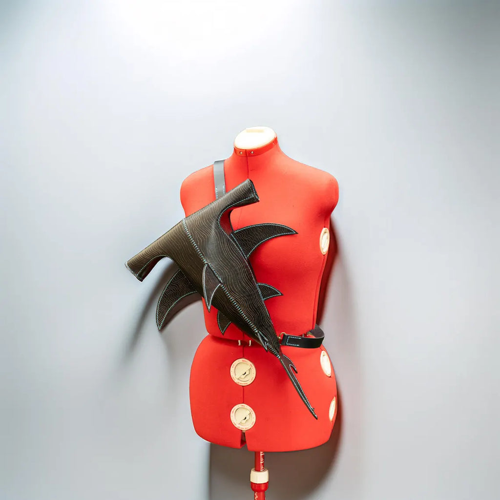 A dark brown leather wing-shaped belt or strap accessory displayed on a red mannequin torso against a light gray background. Made by ATOM Leather.