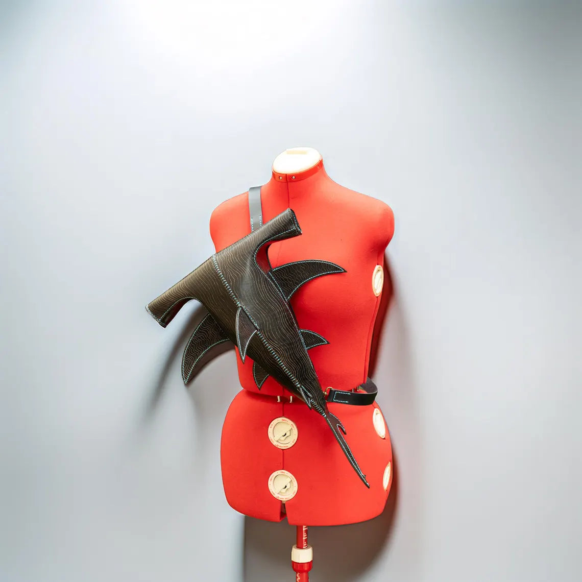A dark brown leather wing-shaped belt or strap accessory displayed on a red mannequin torso against a light gray background. Made by ATOM Leather.