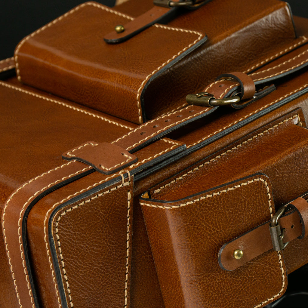 Close-up of a brown leather bag with gold hardware on a dark background.Brown leather Toshiro backpack on a white background, designed and handcrafted by ATOM LEATHER.