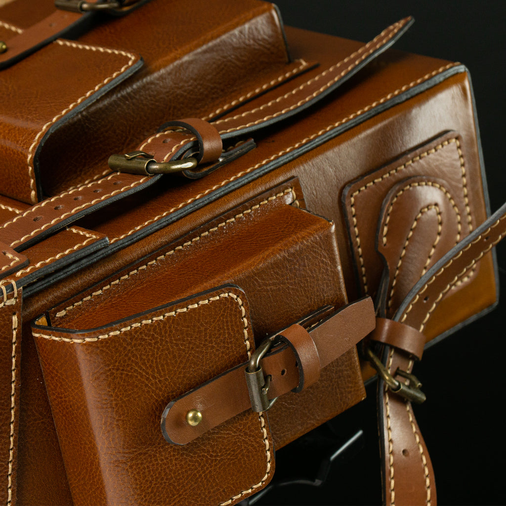 Close-up of a brown leather bag with gold hardware on a black background. Brown leather Toshiro backpack on a white background, designed and handcrafted by ATOM LEATHER.